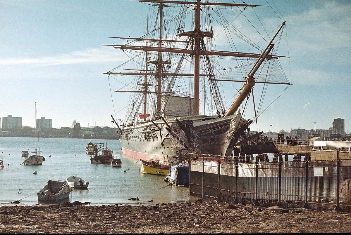A large sailing ship is docked in a harbor with smaller boats in the water and buildings on the distant shore.