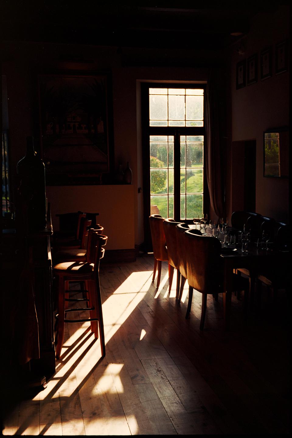 Sunlight streams through a tall window into a dining room with tables and chairs.