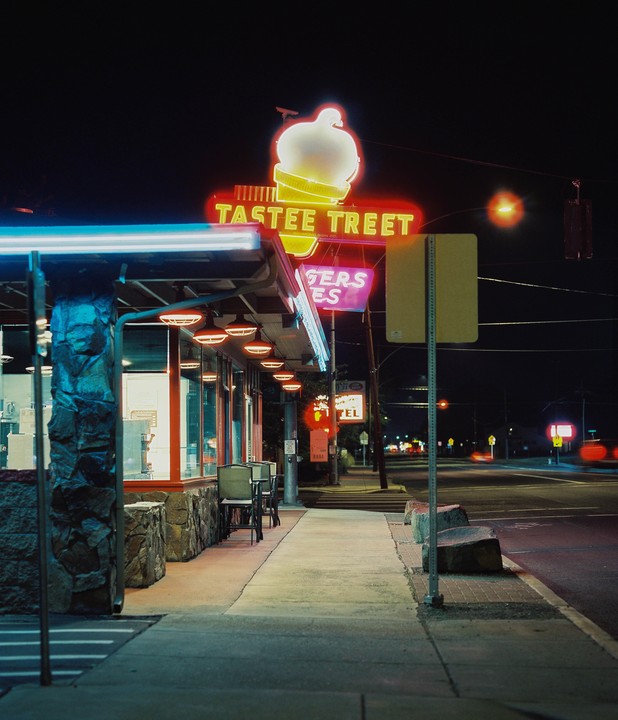 A Tastee Treet ice cream shop with a neon sign is visible at night from a sidewalk next to a street.