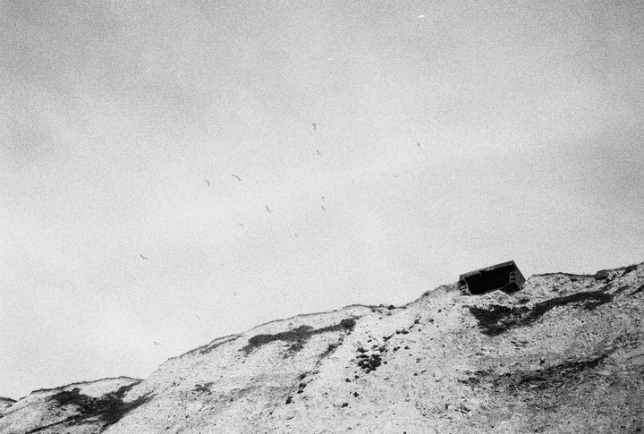 A black and white landscape shows a cliff with a bunker on top and birds flying in the sky.