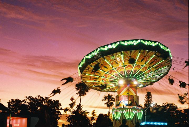 A brightly lit swing ride spins against a vibrant sunset sky with palm trees silhouetted below.