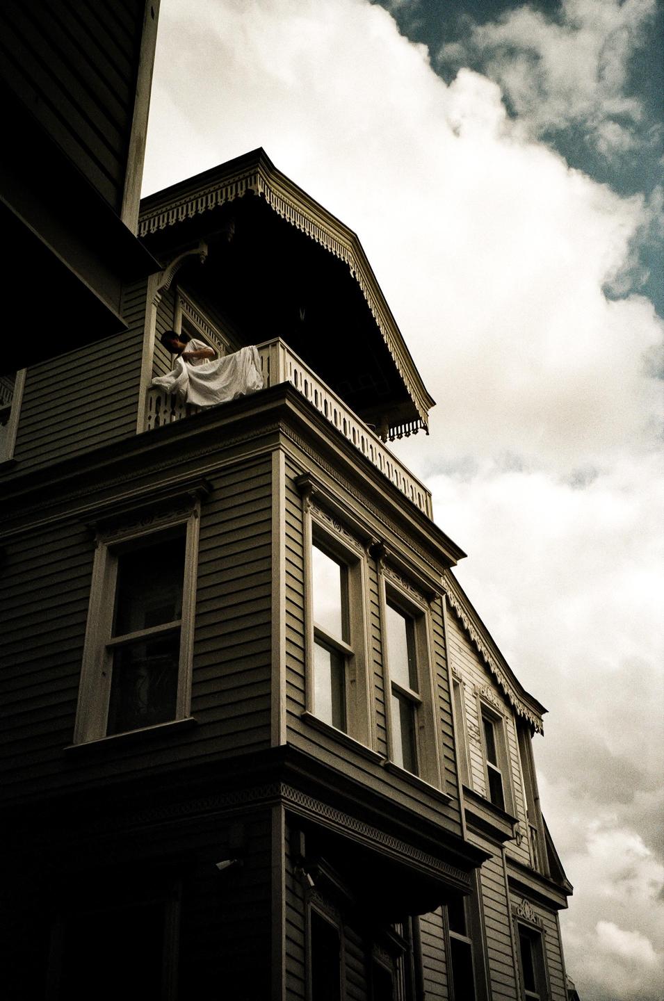 A woman in a white dress leans over a balcony railing on a wooden house under a cloudy sky.