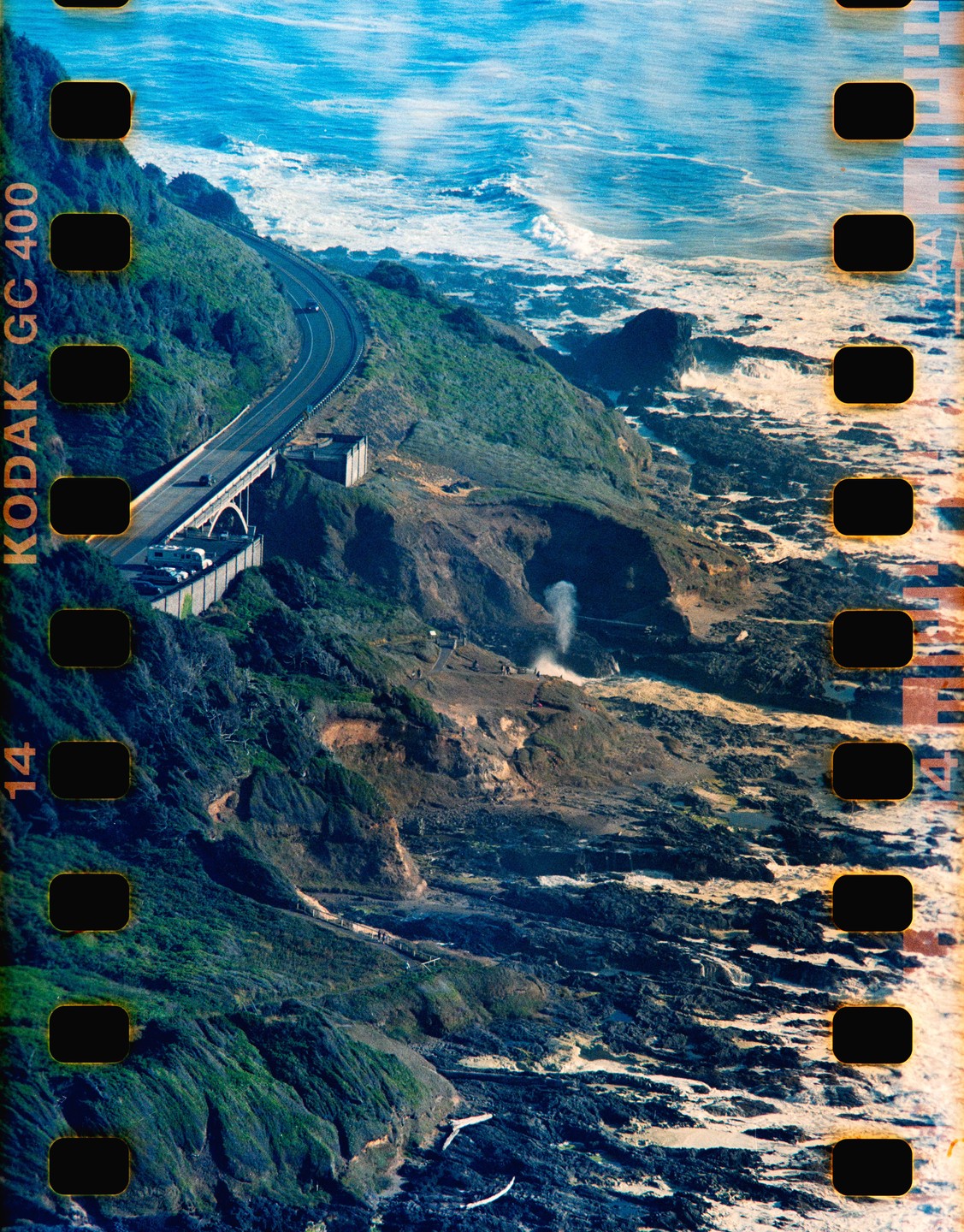 A coastal road winds along a cliff overlooking the ocean, with a geyser erupting from a rock formation.