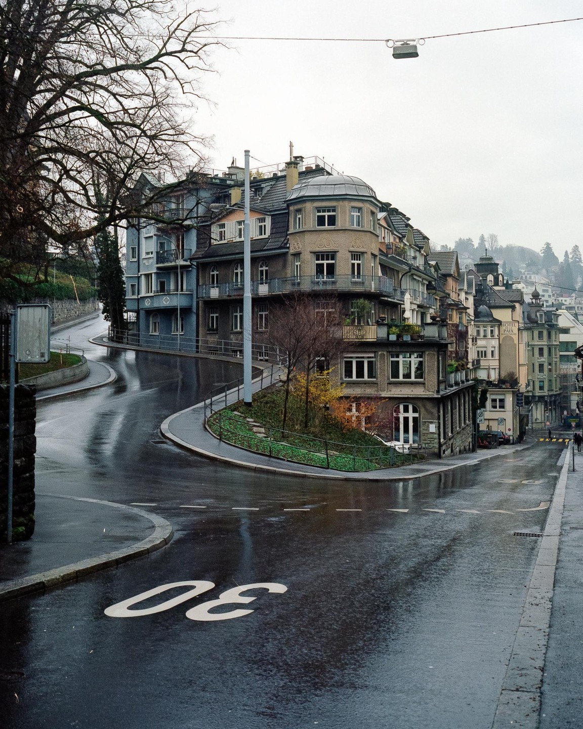A wet street with a painted number 30 on the asphalt curves around a building with multiple windows and balconies.