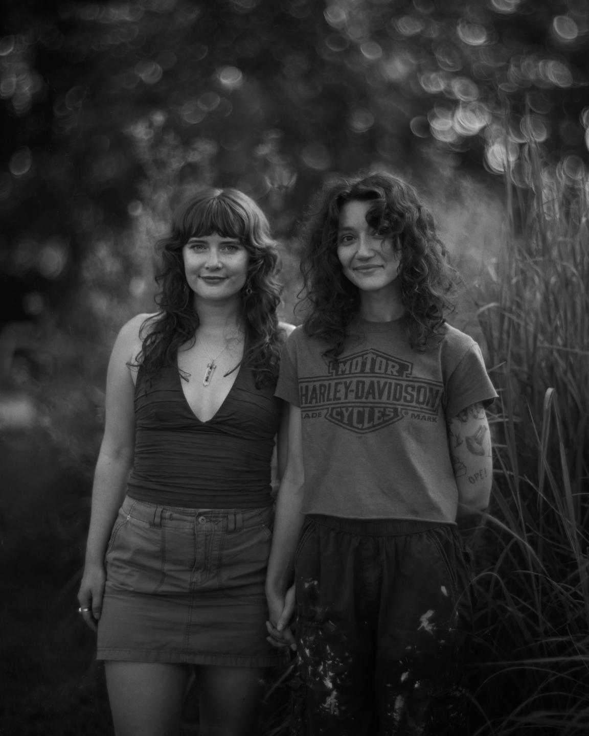 Two women stand side by side, holding hands, with foliage in the background.