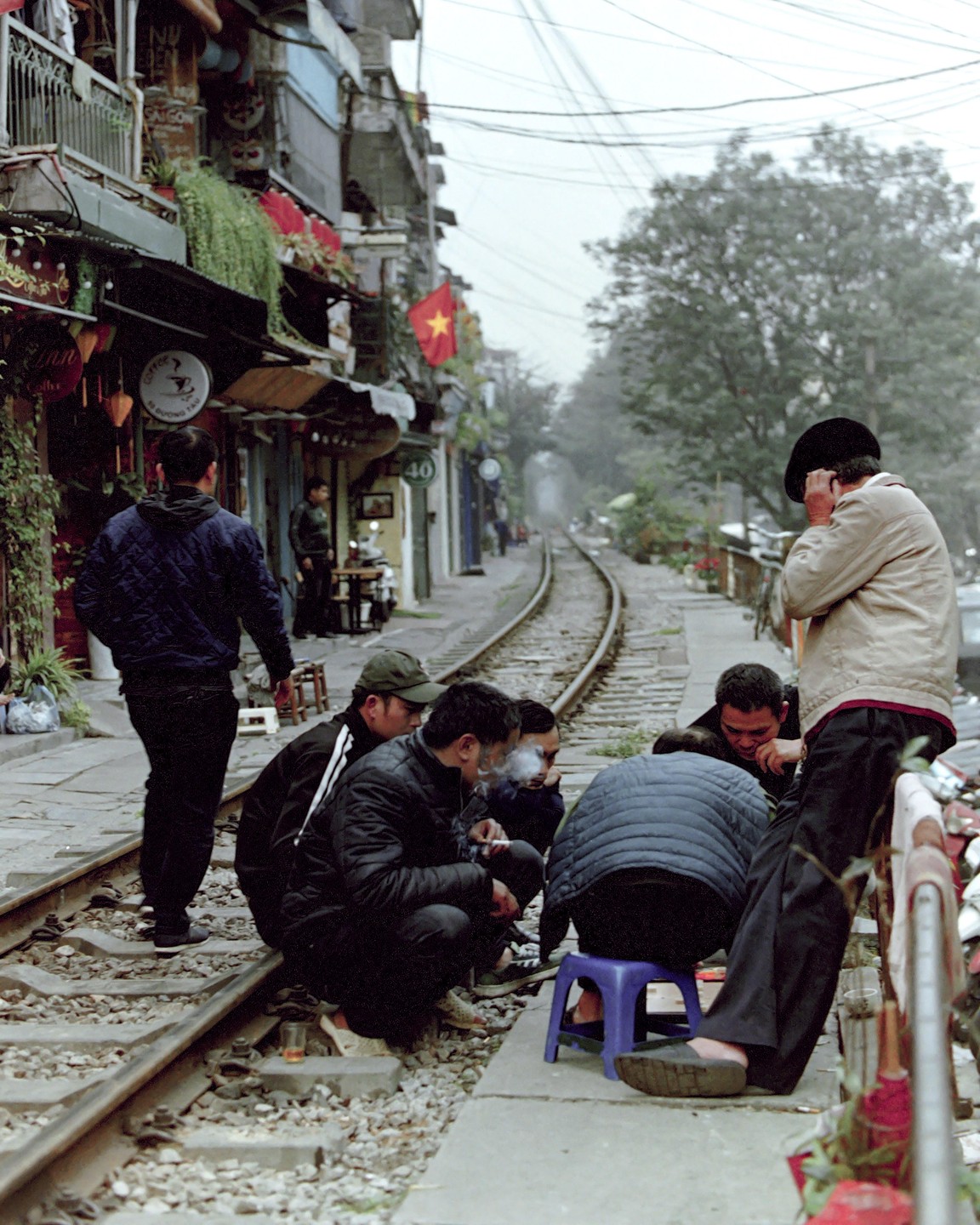 A group of men are gathered on the side of a railway track in a city with buildings lining the tracks.