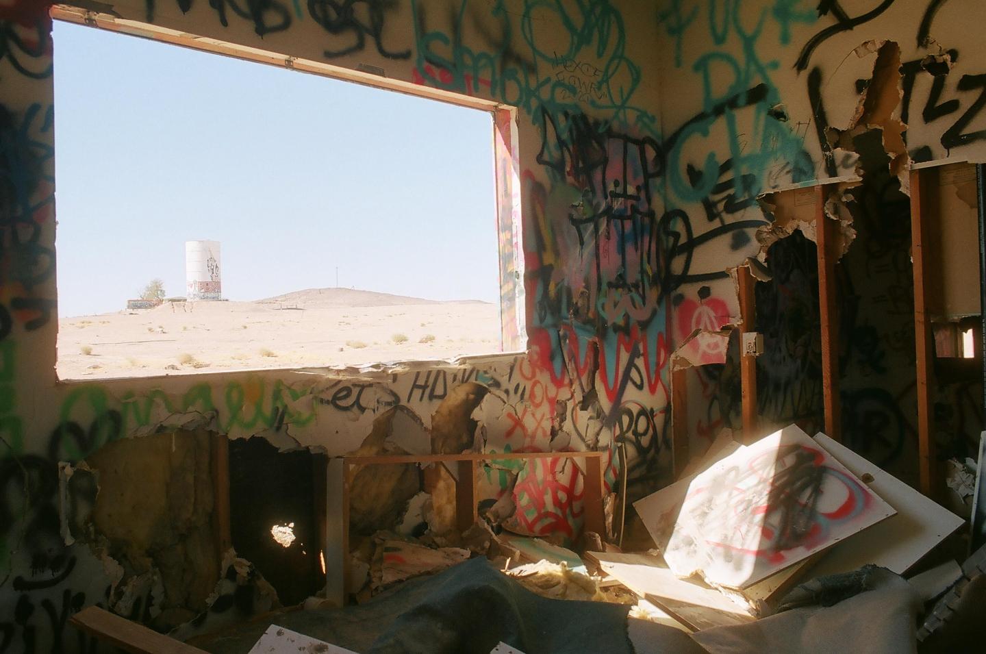 A derelict room covered in graffiti looks out through a window onto a desert landscape with a water tower on a hill.