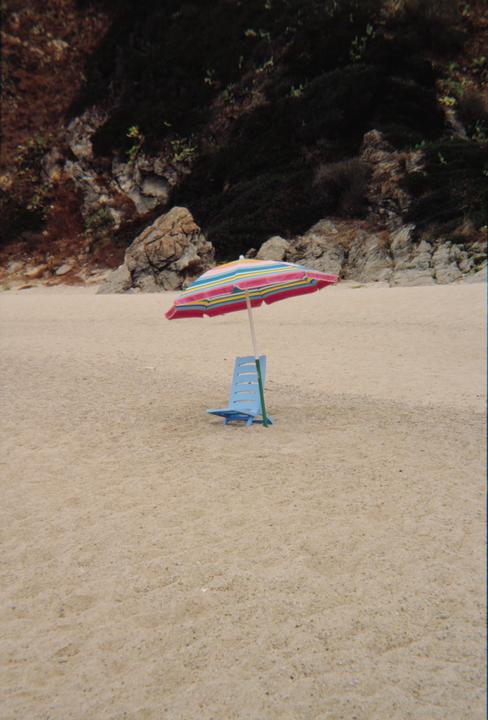 A colorful striped beach umbrella and a blue beach chair sit on a sandy beach with a rocky hill in the background.