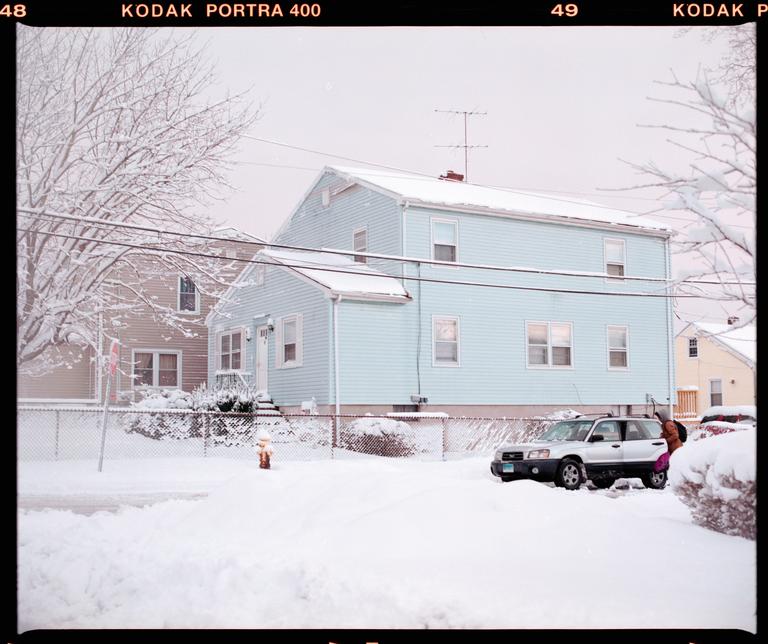 A light blue house and a silver car are covered in snow on a winter day.