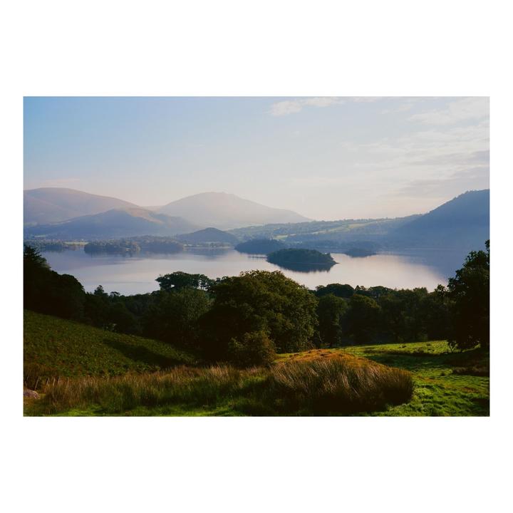 A scenic landscape shows a lake with small islands surrounded by rolling hills and mountains under a hazy sky.