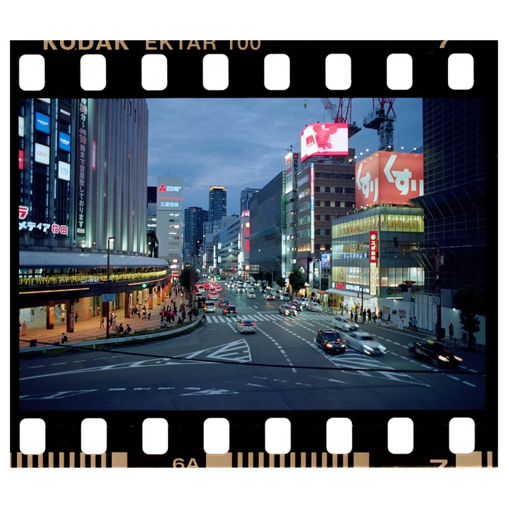 A busy city street at dusk is filled with cars and pedestrians, with tall buildings and bright signs lining the road.