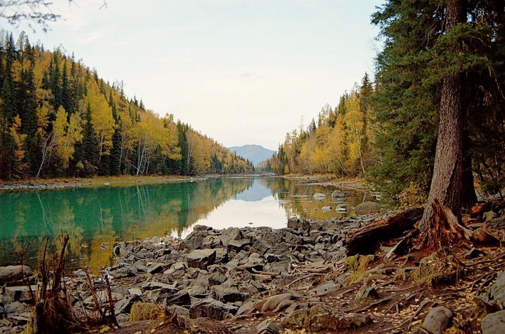 A lake with turquoise water is surrounded by a rocky shore and a forest with autumn foliage.