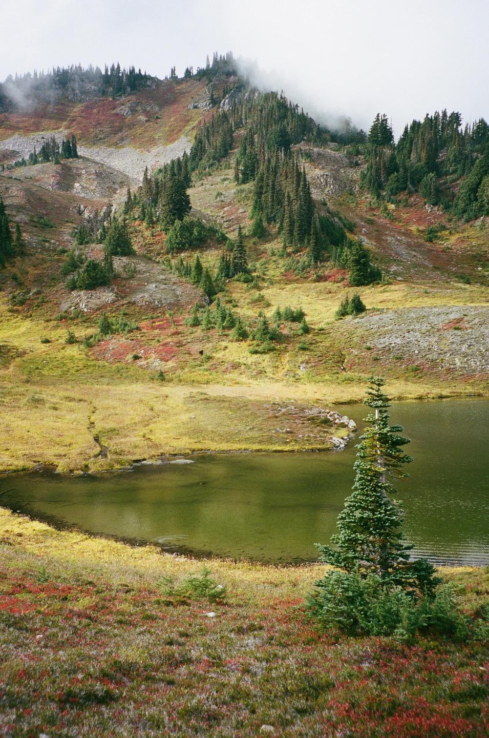 A small lake sits at the base of a mountain covered in trees and colorful autumn foliage under a hazy sky.