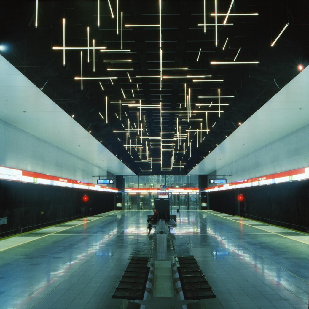 A person sits on a bench in a subway station with a ceiling covered in linear lights.