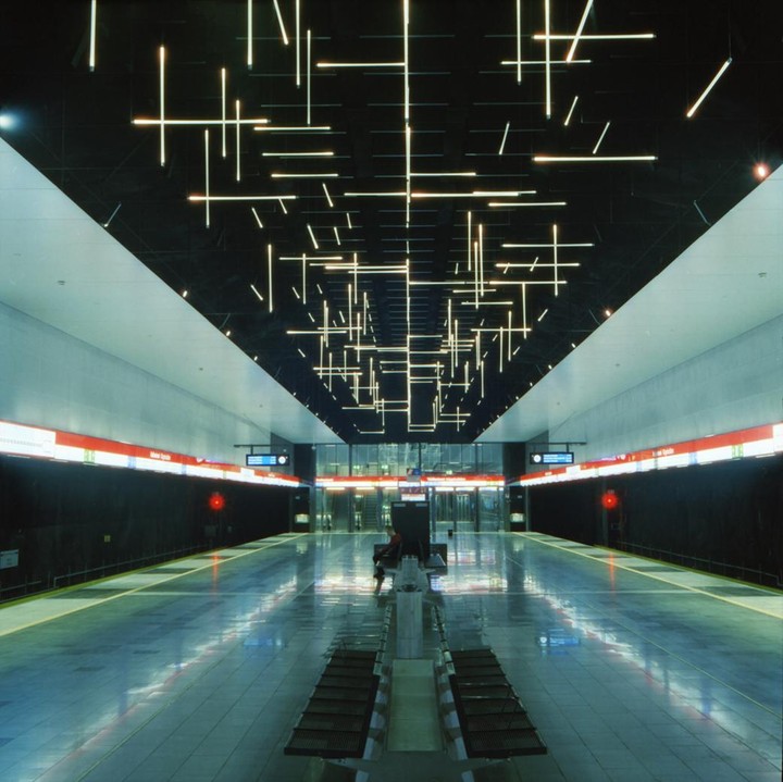 A person sits on a bench in a subway station with a ceiling covered in linear lights.