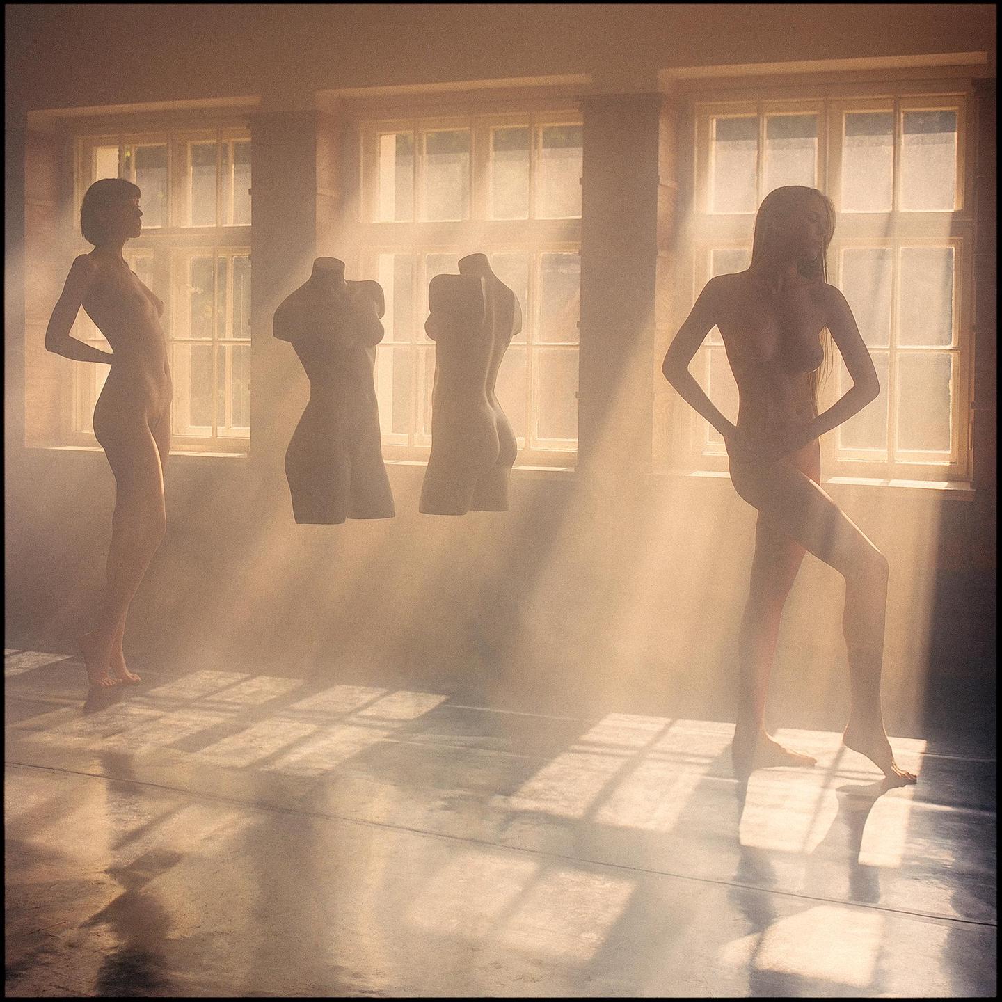 Two nude women and two headless mannequins stand in a hazy room with sunlight streaming through windows.
