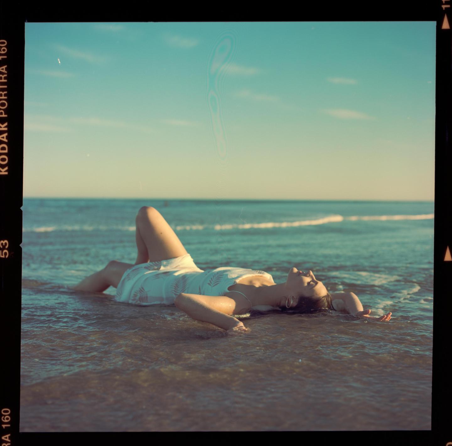 A woman in a white dress lies in the shallow water at the beach.