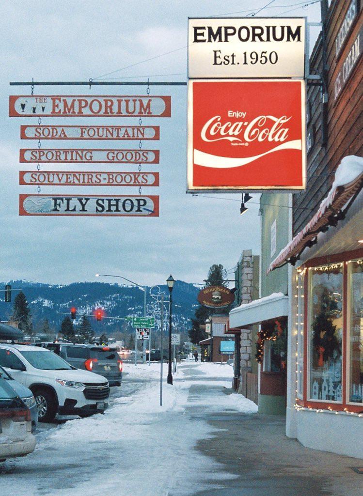 A snow-covered street with cars parked on the side and storefronts with signs, including one for "The Emporium" and a Coca-Cola sign, is visible with mountains in the background.