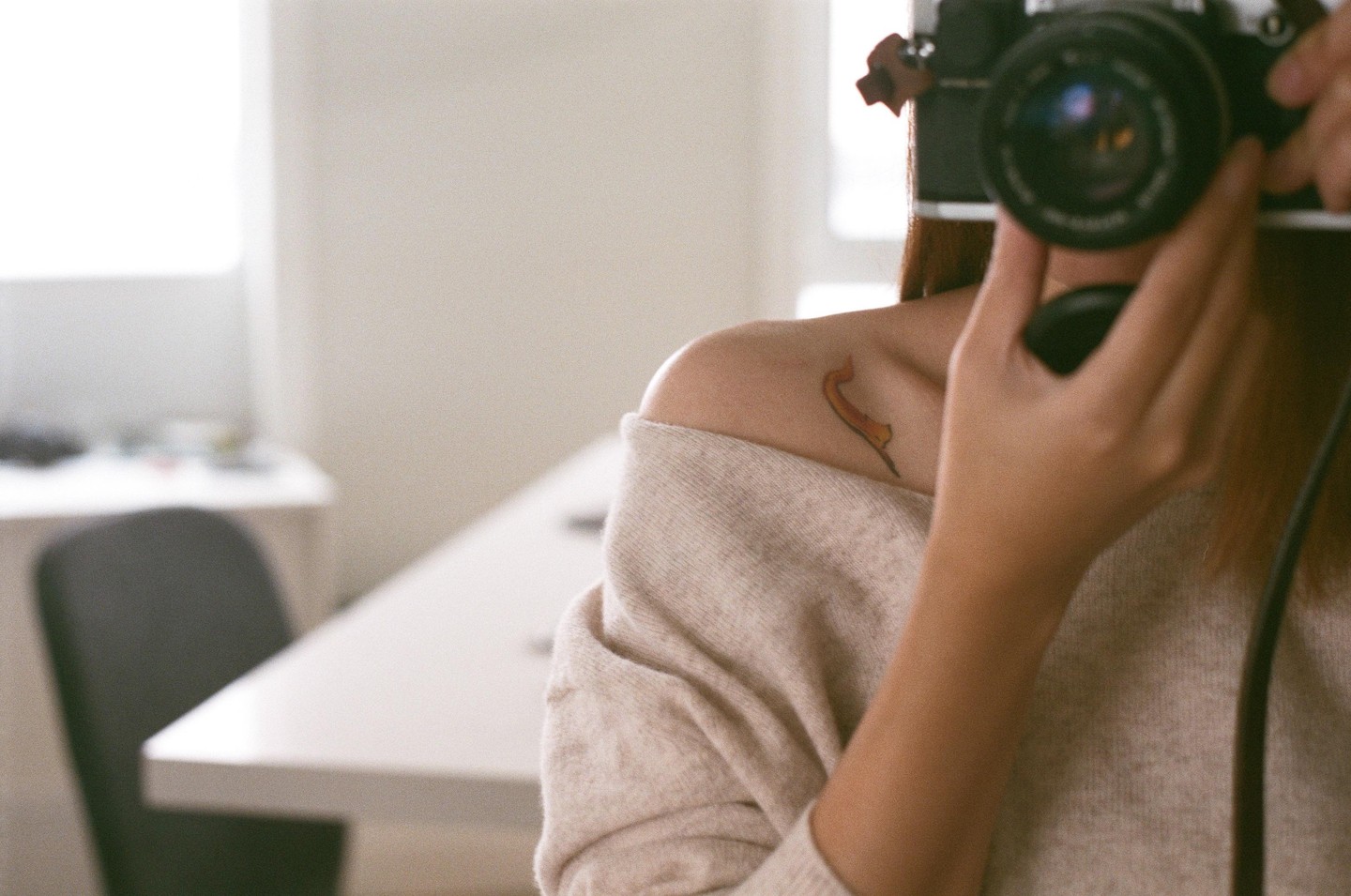 A woman takes a mirror self-portrait, holding a camera to her face, revealing a tattoo on her shoulder.