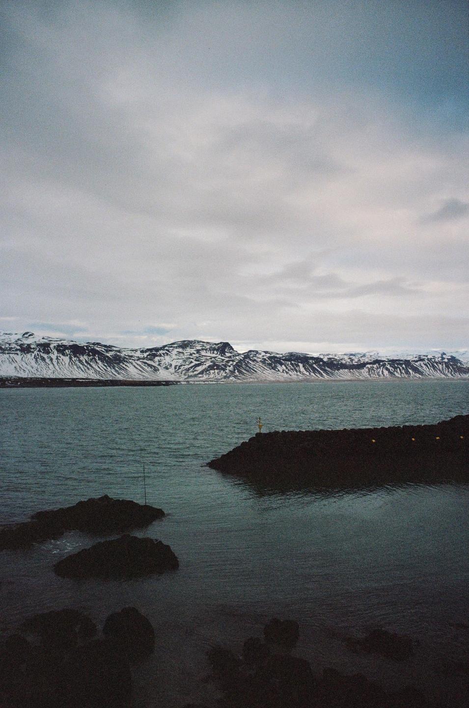 A body of water is in the foreground with snow-capped mountains in the background under a cloudy sky.
