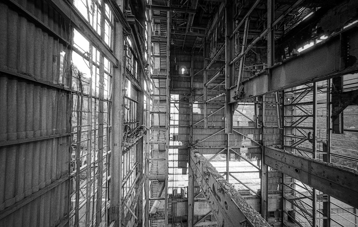 A tall, abandoned industrial building with many windows and metal beams is shown in black and white.