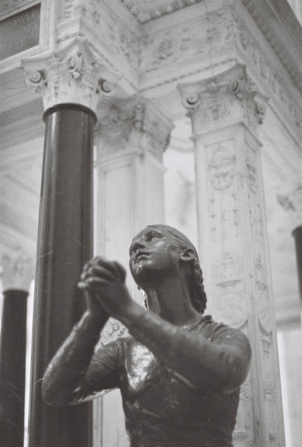 A statue of a woman with clasped hands looks up towards a decorative ceiling and columns.