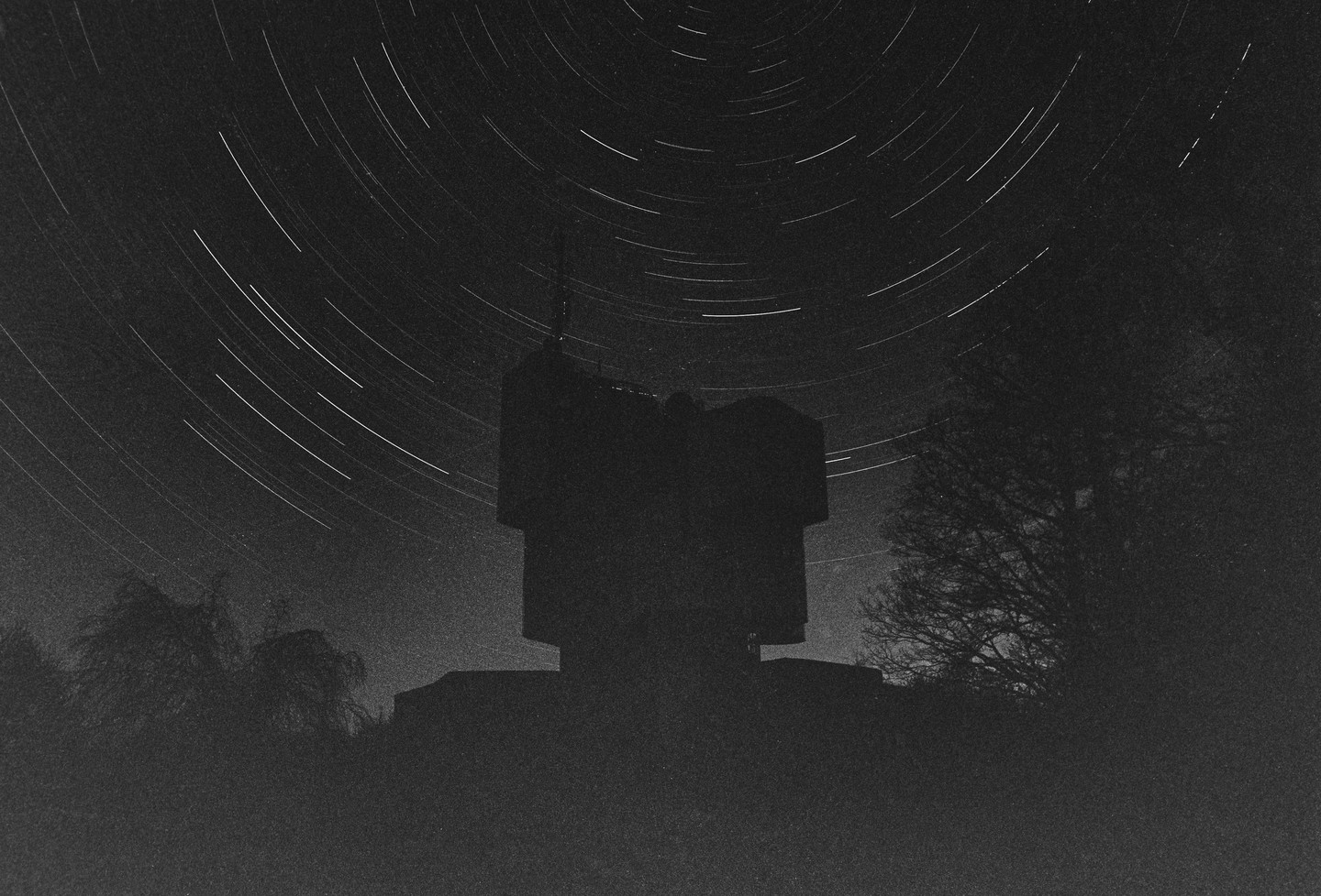 Star trails arc over a dark monument and trees at night.