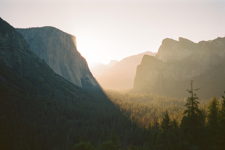 Sunlight illuminates a valley with a forest and mountains, casting a long shadow across the trees.