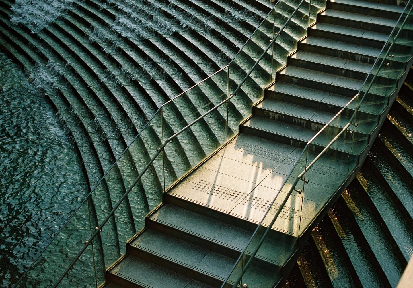 A modern staircase with a glass railing descends alongside a water feature with cascading steps.