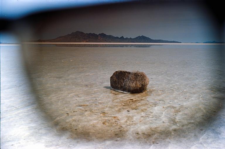 A single rock sits in shallow water with mountains in the distance, viewed through a dark, blurry foreground.