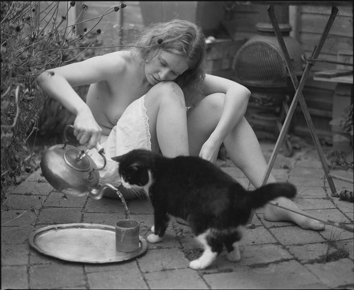 A woman pours water from a kettle into a cup for a cat on a brick patio.