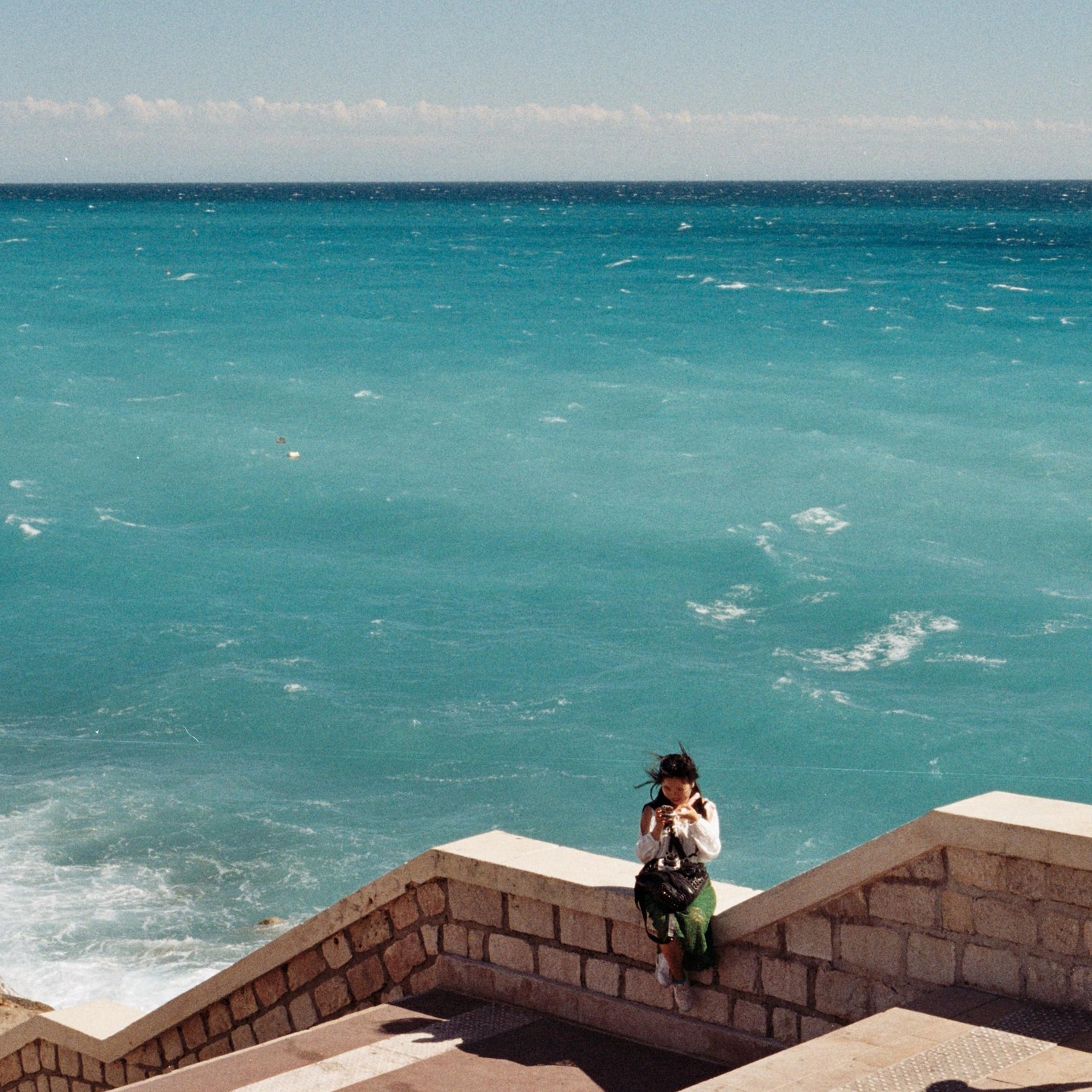 A woman sits on a stone wall overlooking the turquoise sea.