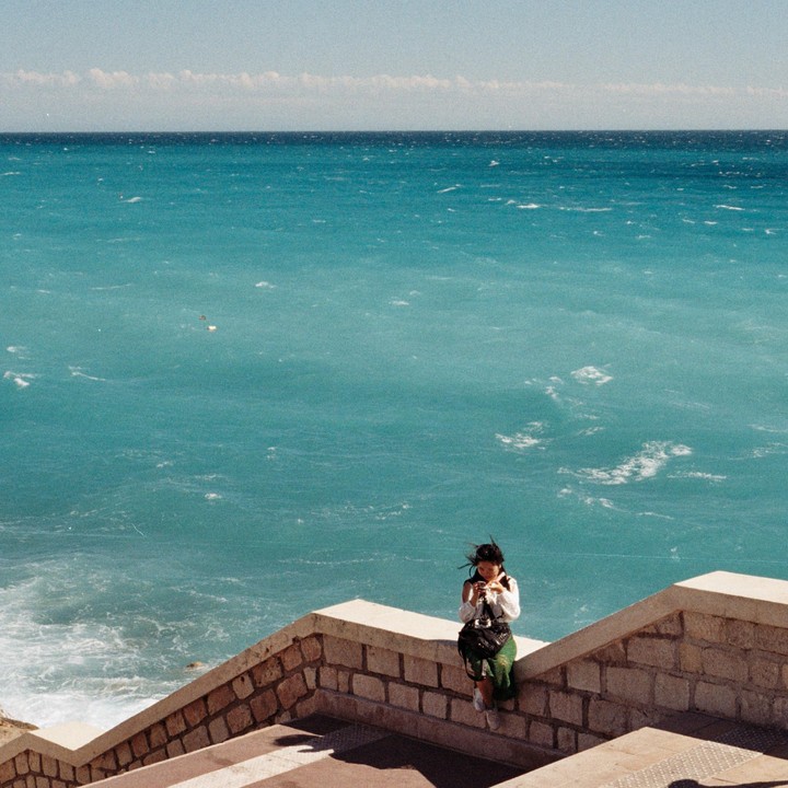 A woman sits on a stone wall overlooking the turquoise sea.