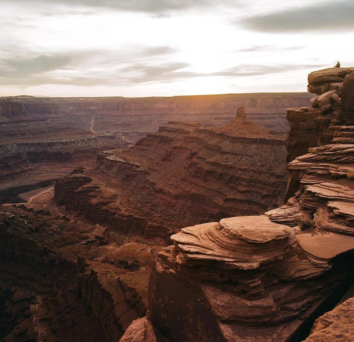 A person sits on a cliff overlooking a vast canyon landscape under a cloudy sky with a hint of sunset.