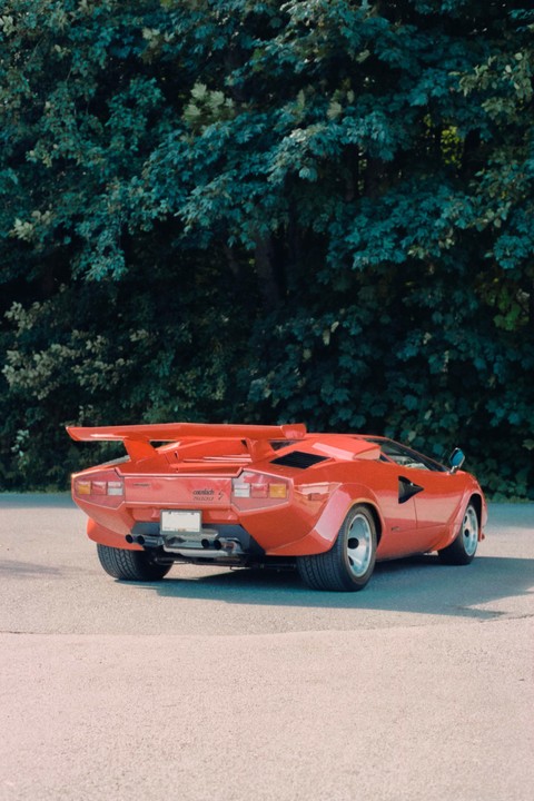 A red Lamborghini Countach is parked on asphalt with green foliage in the background.