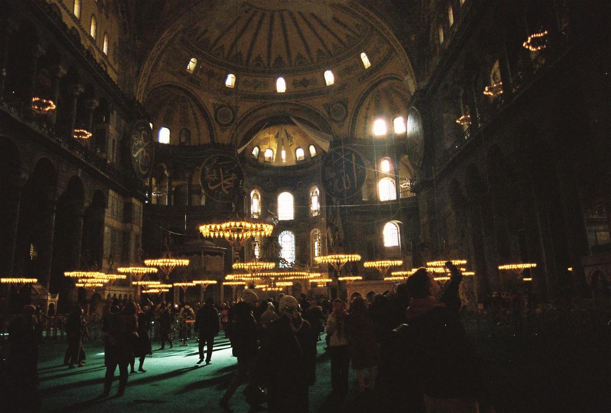 A large crowd of people stands under many chandeliers in a vast, dimly lit hall with high arched ceilings and windows.