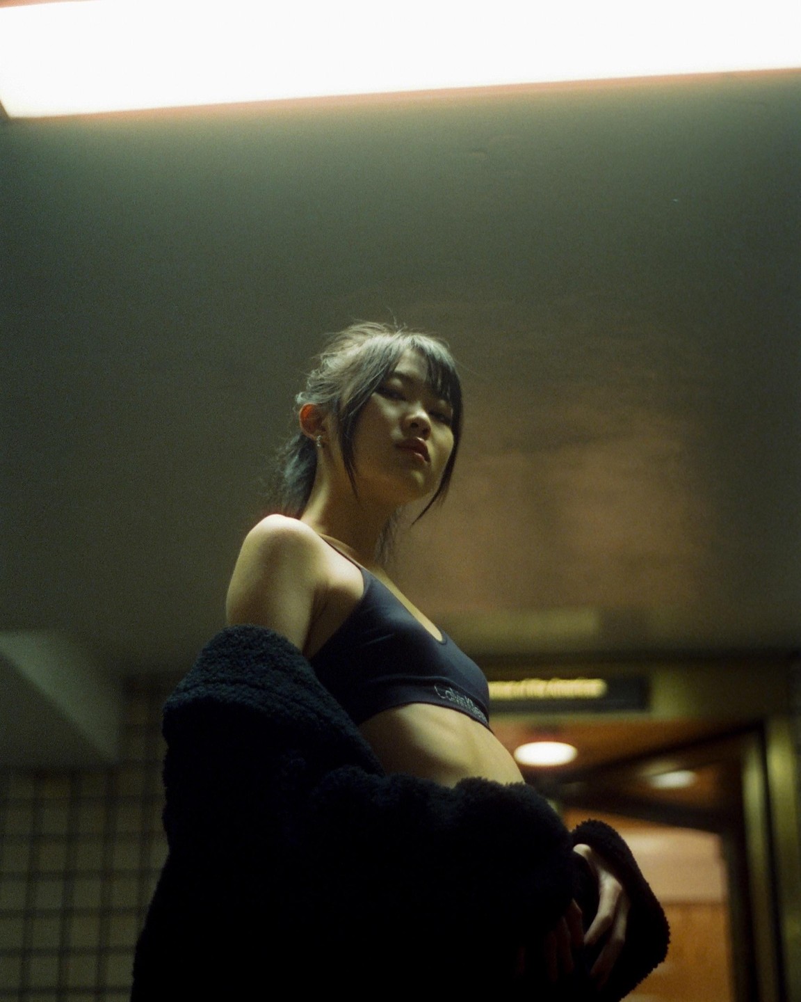 A woman in a sports bra and a dark fluffy jacket looks up at the camera from a low angle.