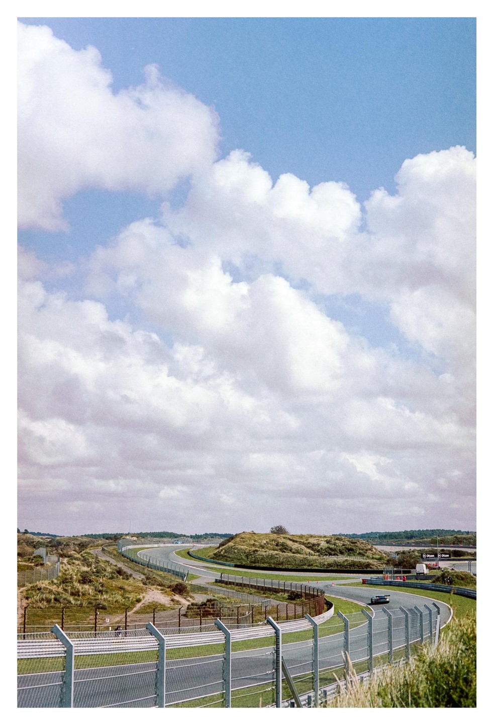 A race track curves through a landscape of grassy dunes under a sky filled with clouds.