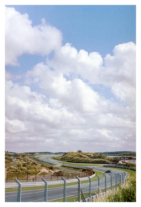 A race track curves through a landscape of grassy dunes under a sky filled with clouds.