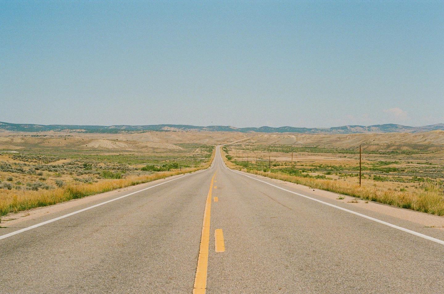 A long, straight road with a yellow dividing line extends into the distance through a dry, grassy landscape under a clear sky.