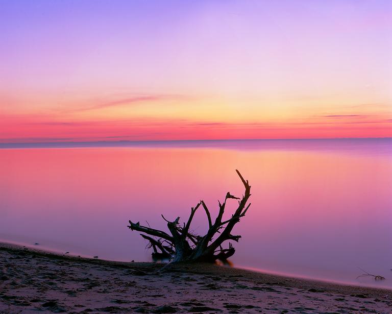 A piece of driftwood rests on a sandy beach with a colorful sky and calm water in the background.