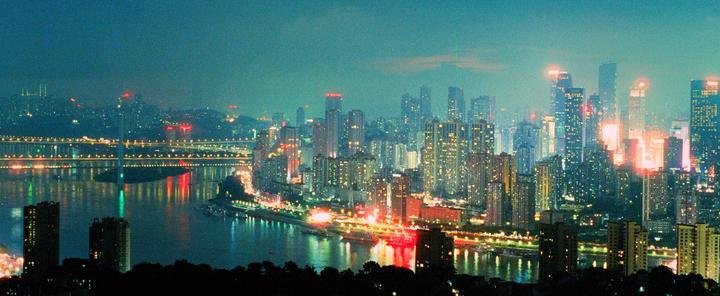 A panoramic night cityscape shows a river with bridges and illuminated buildings reflecting in the water.
