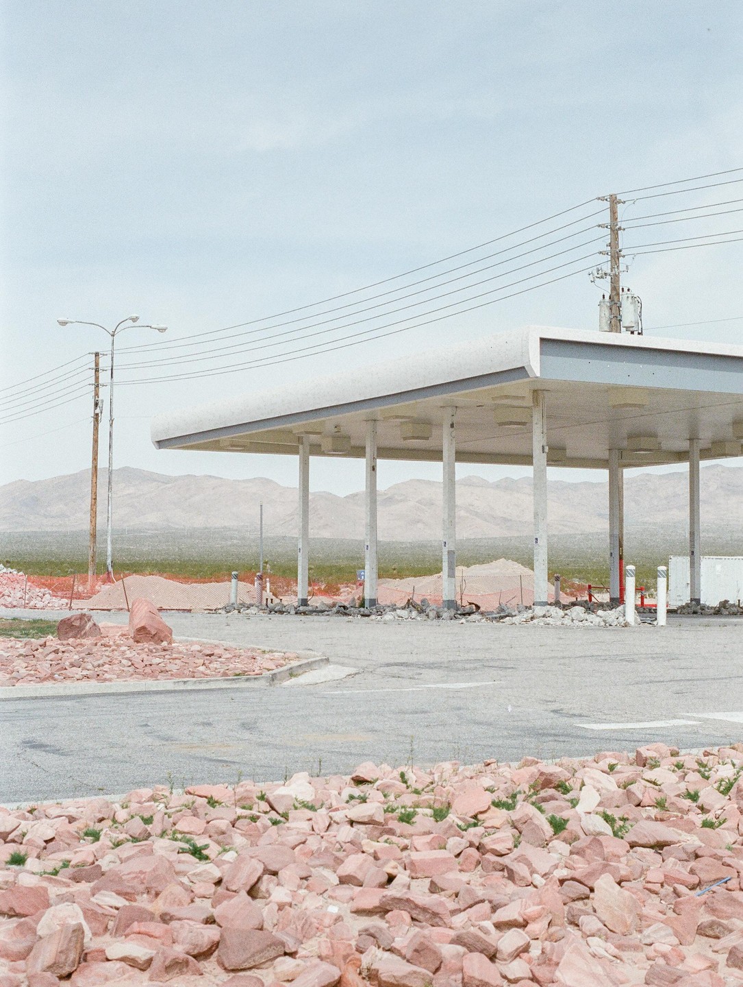 An abandoned gas station with a white canopy stands amidst a rocky, desert landscape under a pale sky.