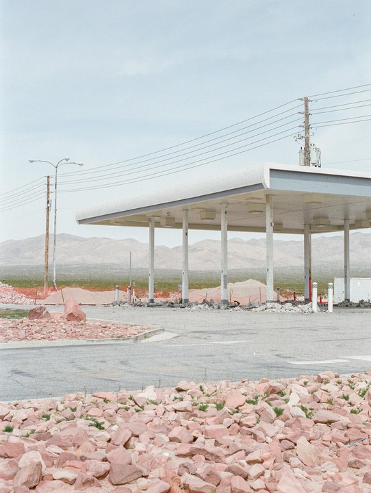 An abandoned gas station with a white canopy stands amidst a rocky, desert landscape under a pale sky.