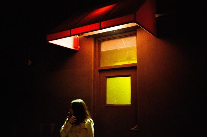 A woman stands in the dark next to a door with yellow windows, illuminated by a red awning.
