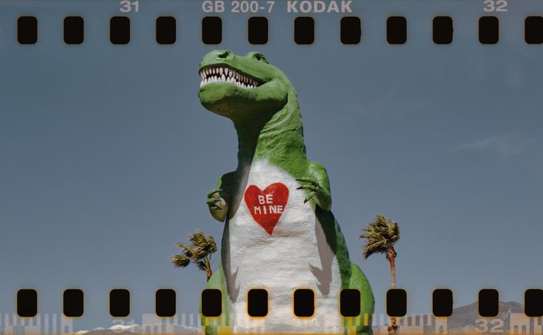 A large green dinosaur statue with a white belly and a red heart that says "Be Mine" stands against a clear sky with palm trees.