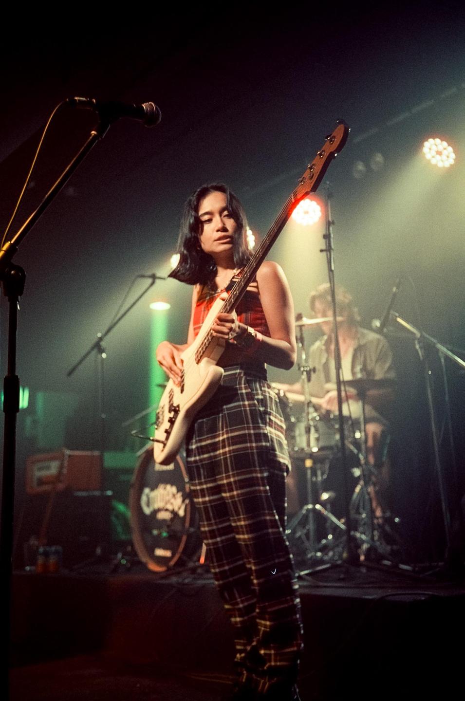 A woman plays a bass guitar on a stage with a drummer and microphone in the background.