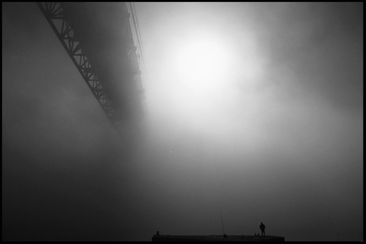A man stands on a pier under a bridge in thick fog with a bright light source in the distance.