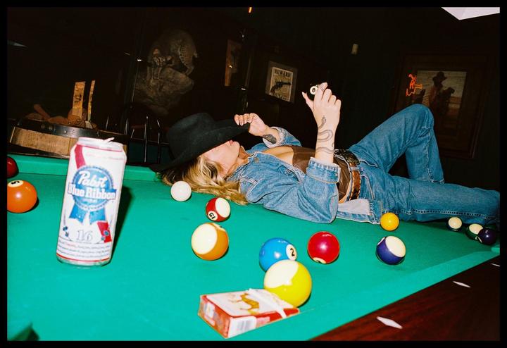 A woman wearing a cowboy hat and denim jacket lies on a pool table surrounded by billiard balls and a beer can.