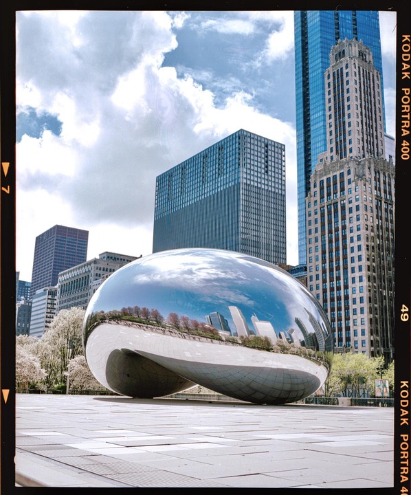 A large, reflective bean-shaped sculpture sits on a paved plaza with tall buildings and a cloudy sky in the background.